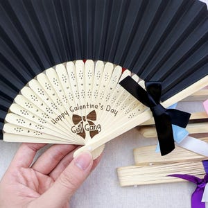 May include: A black folding hand fan with a light-colored wooden frame featuring laser-engraved text that reads Happy Galentine's Day and Girl Gang with a bow icon. The fan is accented with a black ribbon bow and includes additional fans with pink, white, and purple ribbons.