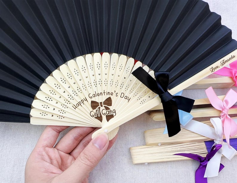 May include: A black folding hand fan with a light-colored wooden frame featuring laser-engraved text that reads Happy Galentine's Day and Girl Gang with a bow icon. The fan is accented with a black ribbon bow and includes additional fans with pink, white, and purple ribbons.