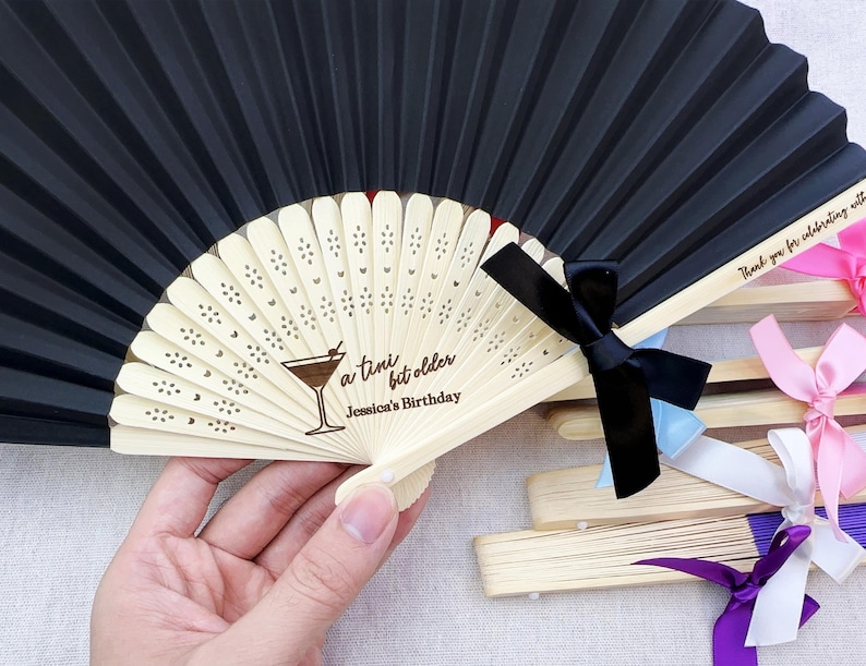 May include: Black paper folding fan with a white wooden handle and a personalised message that reads "a tiny bit older Jessica's Birthday". The fan has a black ribbon tied around the handle.