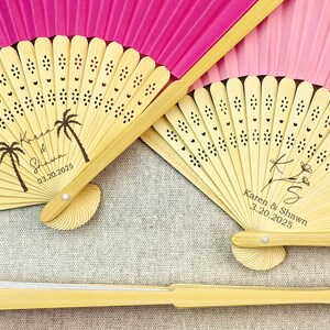 May include: Two personalized bamboo hand fans with pink paper. One fan has a palm tree design and the text "Karen & Shawn 03.20.2025". The other fan has a floral design and the text "Karen & Shawn 3.20.2025".