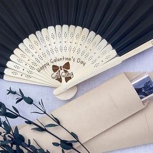 May include: A black folding hand fan with a light-colored wooden frame featuring laser-engraved text that reads Happy Galentine's Day Girl Gang and Love, Amanda. The fan is accompanied by a light brown paper sleeve and a decorative dark green branch.