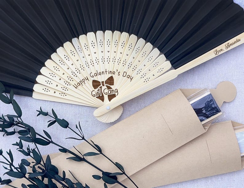 May include: A black folding hand fan with a light-colored wooden frame featuring laser-engraved text that reads Happy Galentine's Day Girl Gang and Love, Amanda. The fan is accompanied by a light brown paper sleeve and a decorative dark green branch.
