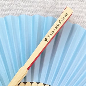 May include: A light blue paper fan with a bamboo handle. The handle has a red edge and the text "Karen's bridal shower" printed on it. The fan is open, displaying its pleated design.