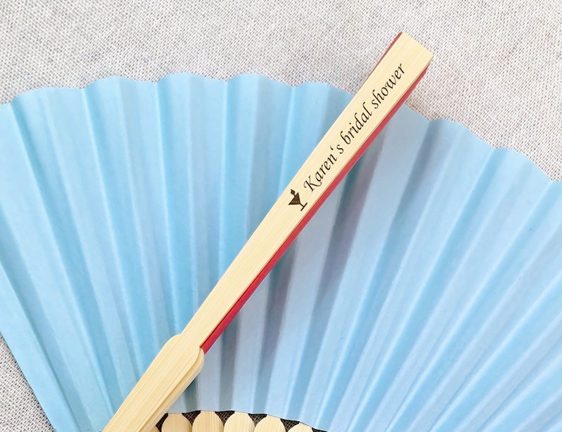 May include: A light blue paper fan with a bamboo handle. The handle has a red edge and the text "Karen's bridal shower" printed on it. The fan is open, displaying its pleated design.