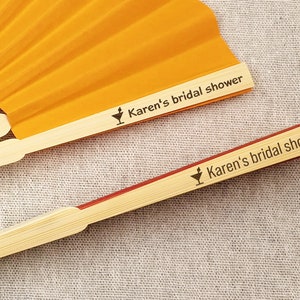 May include: Two open orange paper fans with bamboo handles. The text "Karen's bridal shower" is printed on the handle of each fan, along with a cocktail glass icon. The fans are partially open, displaying the pleated orange paper.