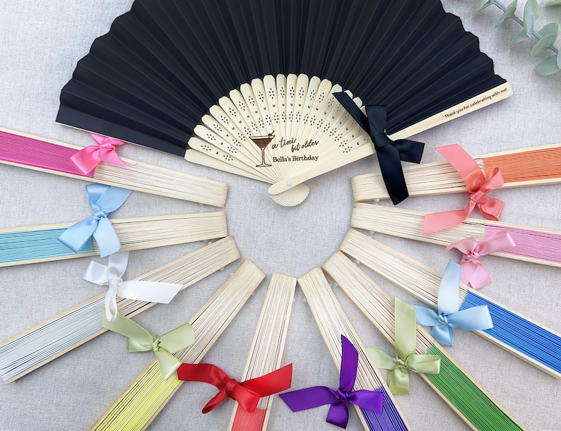May include: A set of 10 black and white paper fans with a wooden handle and a satin ribbon. The fans are decorated with a cocktail glass and the text "a time for bubbles Bella's Birthday".