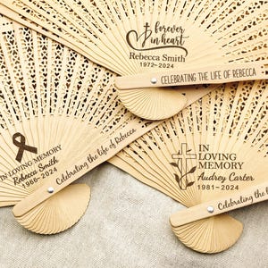 May include: Wooden hand fans with personalised messages for a memorial service. The fans feature a cross, a ribbon, and the text "In Loving Memory" with the name and dates of the deceased.