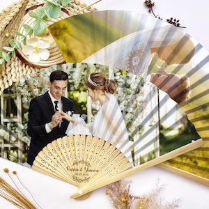 May include: Two wooden hand fans with a wedding photo printed on them. The fans have a light brown color and are decorated with a photo of a couple exchanging rings. The fans are decorated with the names Karen & Simon and the wedding date.