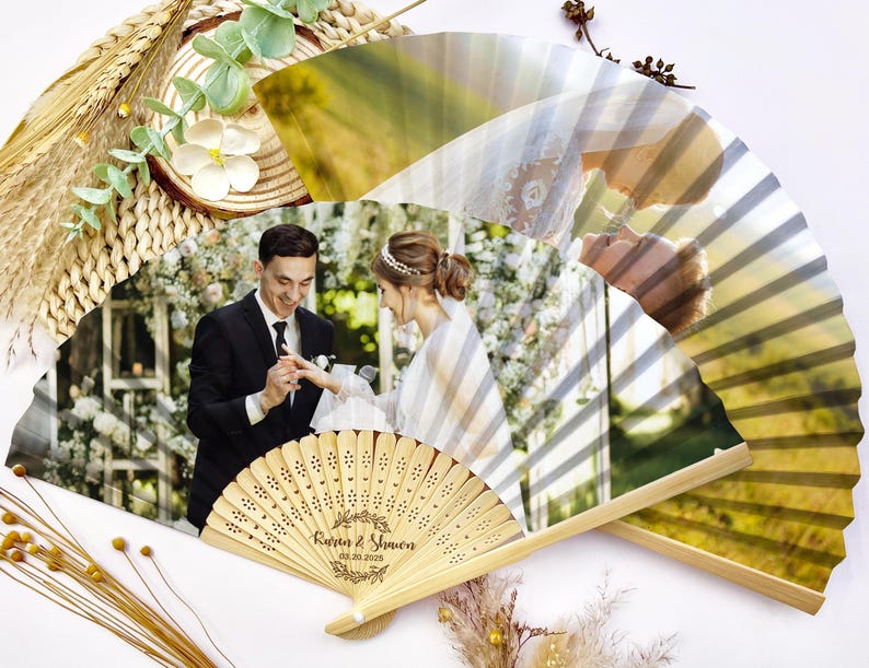May include: Two wooden hand fans with a wedding photo printed on them. The fans have a light brown color and are decorated with a photo of a couple exchanging rings. The fans are decorated with the names Karen & Simon and the wedding date.