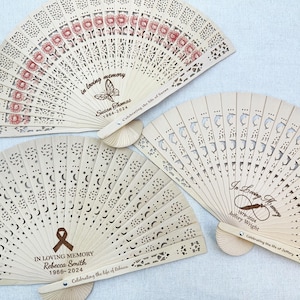 May include: Three wooden hand fans with intricate cutouts and personalized messages. The fans are decorated with floral, star, and dolphin designs. The messages include "In loving memory", "Celebrating the life of", and the names and dates of the deceased.