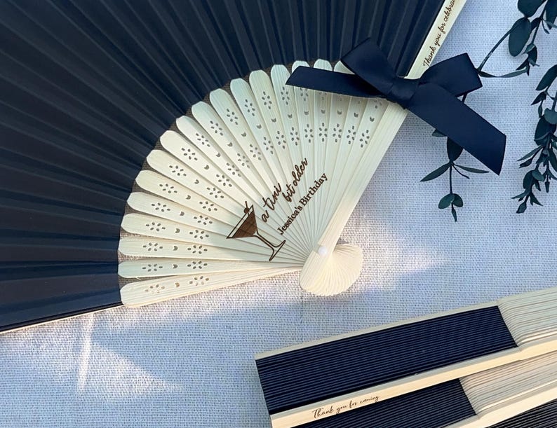 May include: Black and white folding fan with a black ribbon tied around the handle. The fan is decorated with a small cocktail glass and the text "a little bit older" and "Jenica's Birthday".
