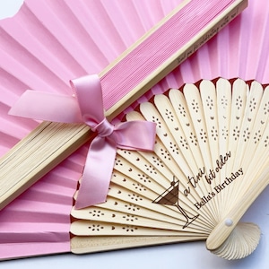 Könnte beinhalten: Ein rosa Papierfächer und ein Holzfächer mit rosa Schleife. Der Holzfächer hat eine Cocktailgrafik und den Text "a tiny bit older Bella's Birthday". Der geschlossene Fächer hat den Text "Thank you for celebrating with us!"