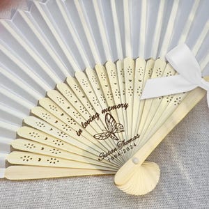 May include: White silk folding fan with a wooden handle. The fan has a butterfly design and the text "In loving memory Susan Thomas 1964-2024" engraved on the handle. The fan has a white ribbon tied around the handle with the text "Celebrating the life" on the ribbon.