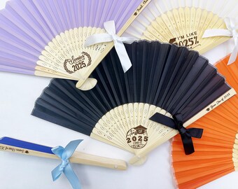 May include: A set of four graduation fans with personalized messages. The fans are made of paper and have a wooden handle. The fans are decorated with a variety of colors, including purple, black, orange, and blue. The fans have different messages, including "Senior Class of 2025", "I'm Like 025%", "Class of 2025 I Did It", and "Frank, Class of 2025".