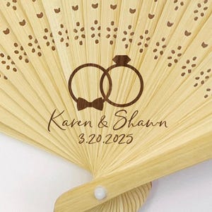 May include: A light-colored bamboo fan with a laser-etched design. The design includes two interlocking rings, a bow tie, and the names "Karen & Shawn" with the date "3.20.2025". The fan has decorative cutouts and a white button.