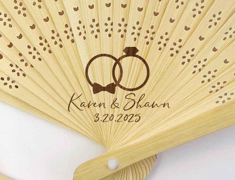 May include: A light-colored bamboo fan with a laser-etched design. The design includes two interlocking rings, a bow tie, and the names "Karen & Shawn" with the date "3.20.2025". The fan has decorative cutouts and a white button.