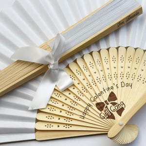 May include: A white folding hand fan with a light wood frame and a white ribbon, paired with a smaller light wood folding fan featuring laser-cut floral patterns, the text "Happy Galentine's Day" and "Girl Gang" with a bow graphic.