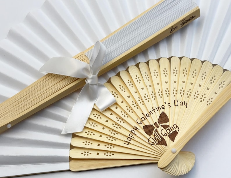 May include: A white folding hand fan with a light wood frame and a white ribbon, paired with a smaller light wood folding fan featuring laser-cut floral patterns, the text "Happy Galentine's Day" and "Girl Gang" with a bow graphic.