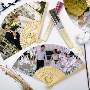 May include: Several personalized wooden fans with wedding photos. The fans feature images of a bride and groom, with intricate laser-cut designs on the wooden handles. Some fans have initials and wedding dates. The fans are arranged on a white surface.