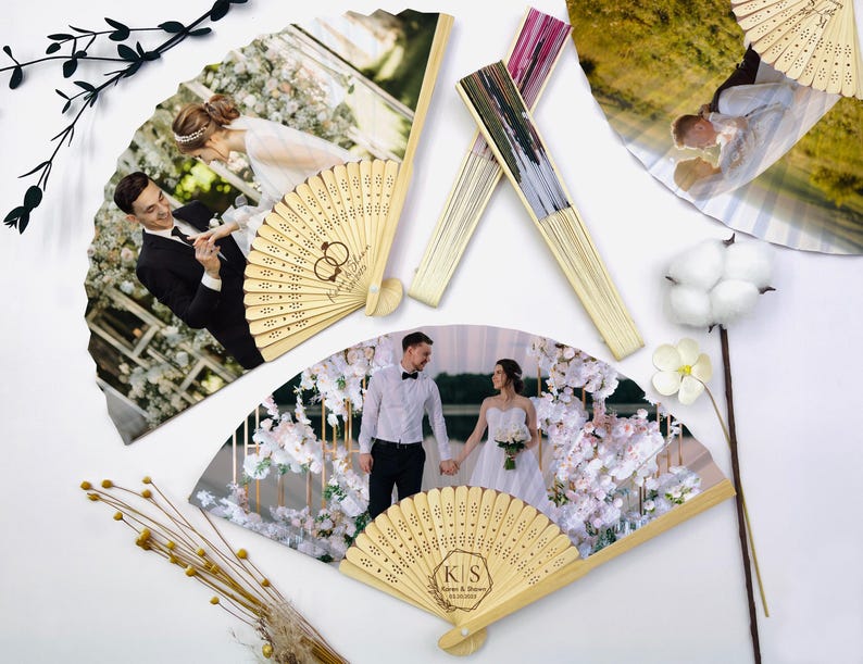 May include: Several personalized wooden fans with wedding photos. The fans feature images of a bride and groom, with intricate laser-cut designs on the wooden handles. Some fans have initials and wedding dates. The fans are arranged on a white surface.