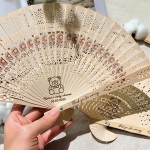 K&ouml;nnte beinhalten: Zwei Holzf&auml;cher mit einem spitzenstickartigen Design und einem Teddyb&auml;r-Motiv. Die F&auml;cher sind mit dem Text "Karen's baby shower 03.20.2025" und "Vielen Dank f&uuml;r Ihr Kommen" personalisiert.