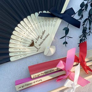 May include: A set of wooden folding fans with black, red, pink, and white ribbons. The fans are decorated with a heart pattern and the text "a tini bit older Bella's Birthday".