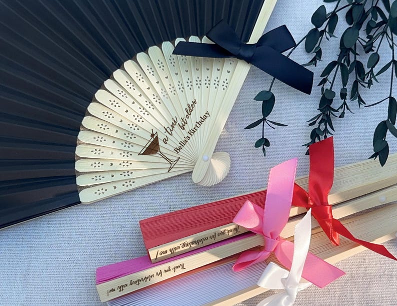 May include: A set of wooden folding fans with black, red, pink, and white ribbons. The fans are decorated with a heart pattern and the text "a tini bit older Bella's Birthday".
