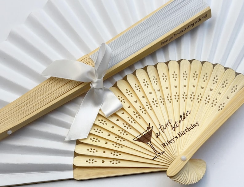 May include: Two white paper fans with wooden handles. One fan has a white satin ribbon tied around the handle. The other fan has a wooden handle with a laser engraved design of a cocktail glass, hearts, and the text "a  tiny bit older Riley's Birthday".
