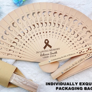 May include: A wooden hand fan with a dolphin design and the text "IN LOVING MEMORY Rebecca Smith 1966-2024 Celebrating the life of Rebecca" engraved on the front. The fan is folded and comes with a brown paper envelope.