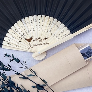 May include: Black folding hand fan with a wooden handle and a personalised message that reads "a tiny bit older Riley's Birthday". The fan is packaged in a brown paper envelope.