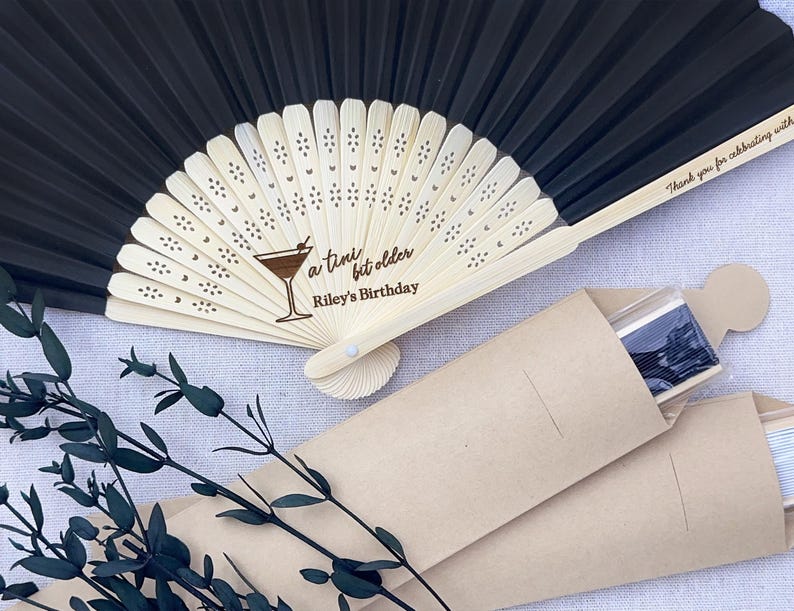 May include: Black folding hand fan with a wooden handle and a personalised message that reads "a tiny bit older Riley's Birthday". The fan is packaged in a brown paper envelope.