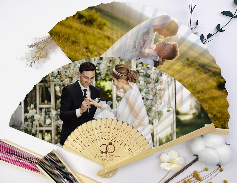 May include: Wooden wedding fan with a photo of a couple exchanging rings. The fan has a laser-cut design and the names "Karen & Shawn" and the date "3-24-2023" are engraved on the handle. The fan is open, displaying a wedding photo.