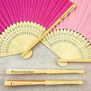 May include: A collection of paper fans in shades of pink and fuchsia, with natural bamboo handles. The fans are open and closed, with the text "Karen's bridal shower" printed on the handles. The fans have a decorative floral pattern.