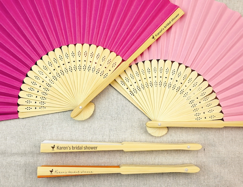 May include: A collection of paper fans in shades of pink and fuchsia, with natural bamboo handles. The fans are open and closed, with the text "Karen's bridal shower" printed on the handles. The fans have a decorative floral pattern.