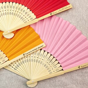 May include: Three colorful paper fans in red, orange, and pink, with natural bamboo handles. Each fan is printed with the text "Karen's bridal shower". The fans are fanned out, showing their pleated design.