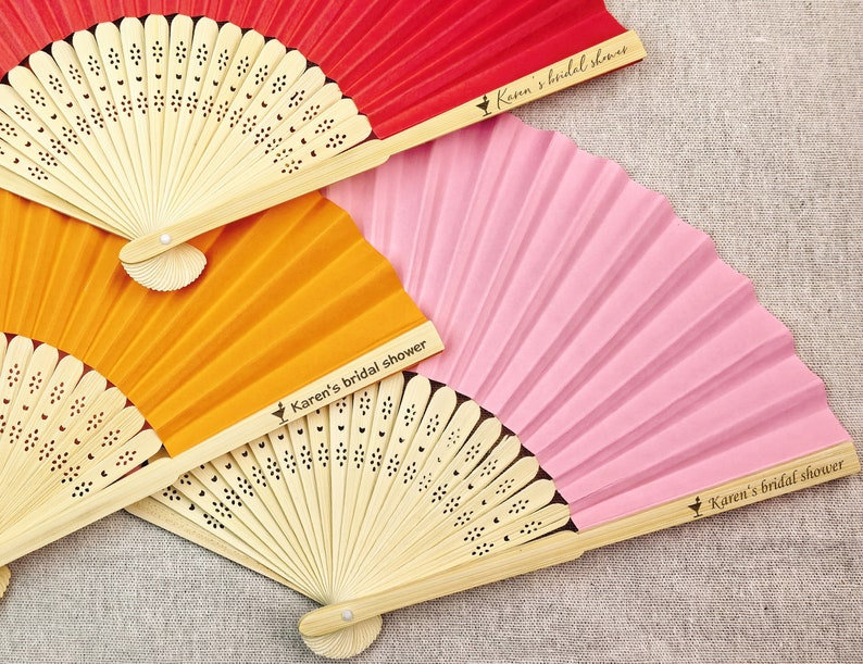 May include: Three colorful paper fans in red, orange, and pink, with natural bamboo handles. Each fan is printed with the text "Karen's bridal shower". The fans are fanned out, showing their pleated design.