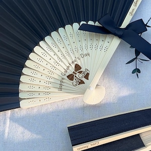 May include: A black folding hand fan with light-colored wooden ribs featuring laser-engraved text that reads Happy Galentine's Day Girl Gang and a brown bow graphic. A black satin ribbon bow is attached to the top, and Love, Amanda is engraved.