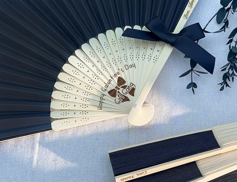 May include: A black folding hand fan with light-colored wooden ribs featuring laser-engraved text that reads Happy Galentine's Day Girl Gang and a brown bow graphic. A black satin ribbon bow is attached to the top, and Love, Amanda is engraved.
