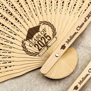 May include: A wooden hand fan with a decorative cut-out design. The fan is personalized with the text "Class of 2025" and the name "Melissa".