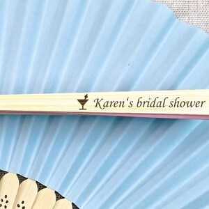 May include: A light blue paper fan with a bamboo handle. The handle is engraved with a cocktail glass icon and the text "Karen's bridal shower". The fan is partially open, revealing its pleated design.