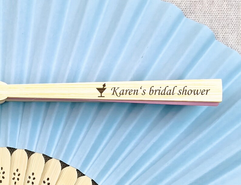 May include: A light blue paper fan with a bamboo handle. The handle is engraved with a cocktail glass icon and the text "Karen's bridal shower". The fan is partially open, revealing its pleated design.