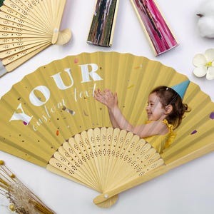 May include: Yellow folding fan with a photo of a child wearing a party hat. The fan has the text "YOUR custom text" in white. The fan is made of wood and paper. Other fans are visible in the background.