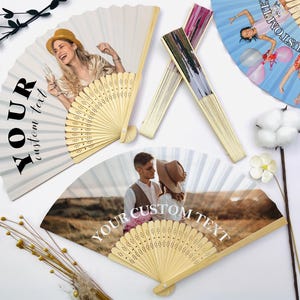 May include: Several open hand fans with custom photos and text. The fans have a light wood frame and a white paper fan. One fan has a photo of a woman holding a champagne glass. Another fan has a photo of a couple. The text "YOUR custom text" is printed on the fans.
