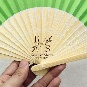 Puede incluir: Un abanico de papel verde y blanco con un marco de bambú. El abanico tiene un diseño floral y las iniciales "K" y "S" con el texto "Karen & Shawn 03.20.2025".