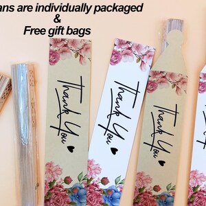 May include: Folded wooden hand fans with intricate cutouts, some in clear plastic sleeves, paired with rectangular paper tags featuring floral illustrations in pink and blue, the text "Thank You" in black script, and a small black heart icon.