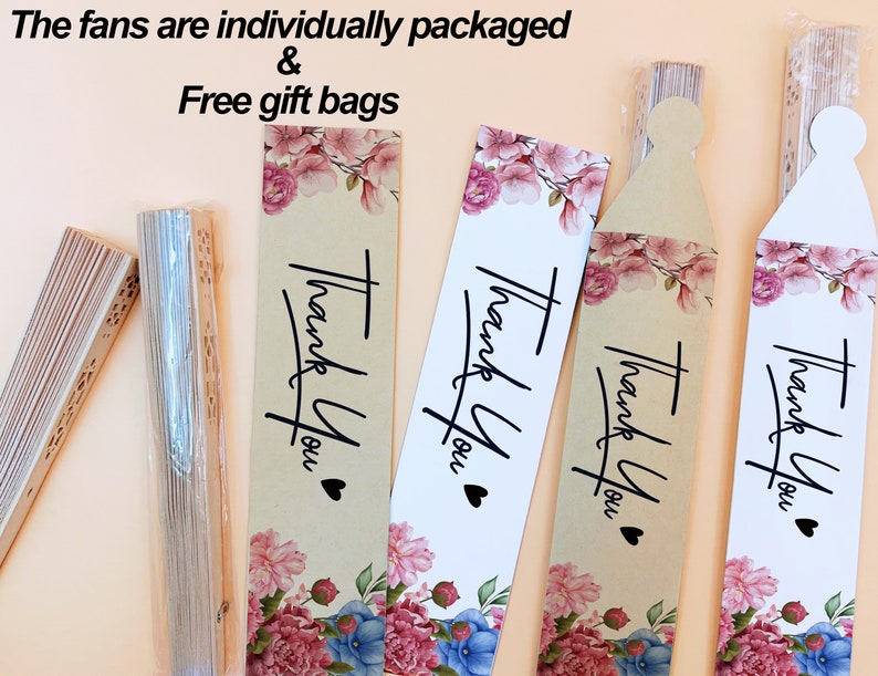 May include: Folded wooden hand fans with intricate cutouts, some in clear plastic sleeves, paired with rectangular paper tags featuring floral illustrations in pink and blue, the text "Thank You" in black script, and a small black heart icon.