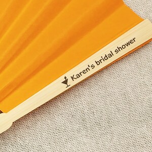 May include: An orange hand fan with a wooden handle. The fan is open, displaying its pleated design. The text "Karen's bridal shower" is printed on the wooden handle, along with a cocktail glass icon.
