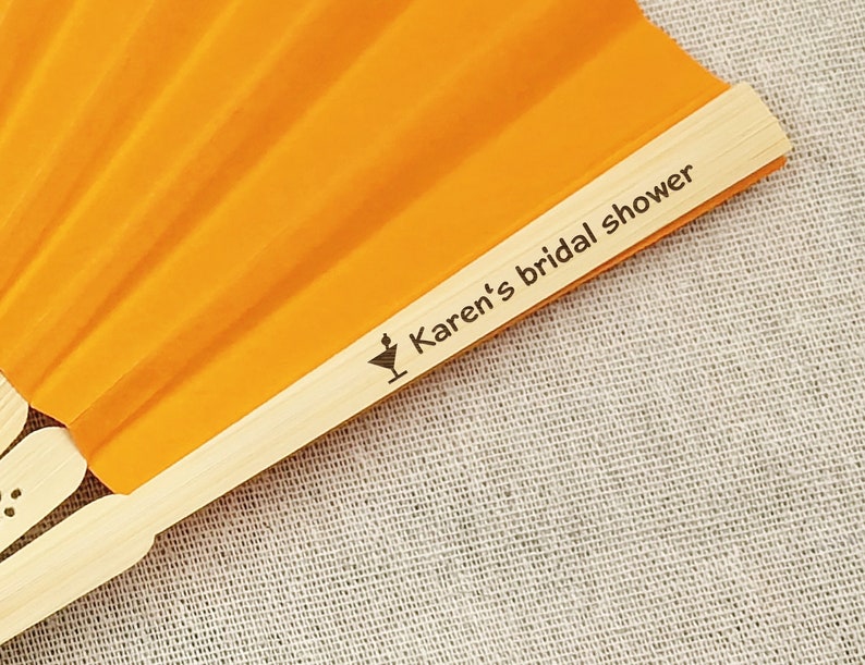 May include: An orange hand fan with a wooden handle. The fan is open, displaying its pleated design. The text "Karen's bridal shower" is printed on the wooden handle, along with a cocktail glass icon.