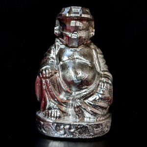 May include: A silver-colored, stylized laughing Buddha statue with a helmet-like head. The statue is sitting with its legs crossed and has a large belly.