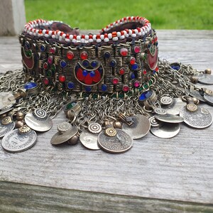 May include: A silver and gold choker necklace with red, blue, and green beads and coin-like charms. The necklace is made of metal and has a beaded band with a decorative design.